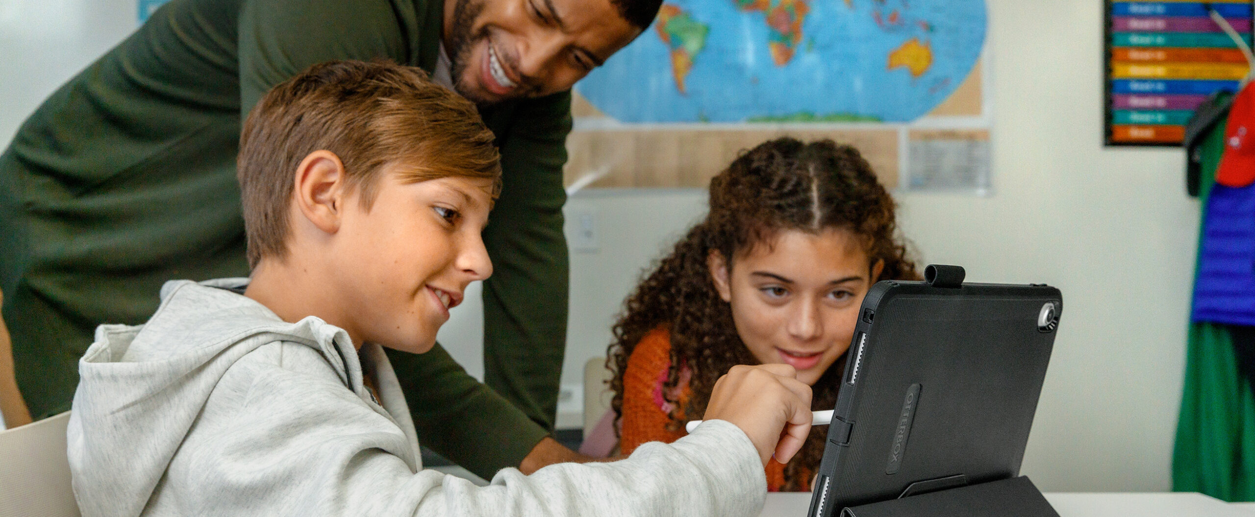 Tablet_Solutions A boy and girl in a classroom learning from a tablet while a teacher overlooks the two